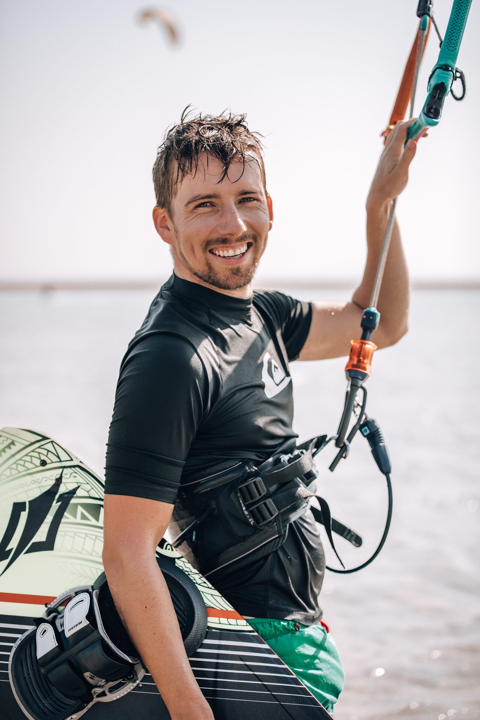 A picture of me smiling at the camera controling the kite with one hand on the bar, and another hand holding a kiteboard.